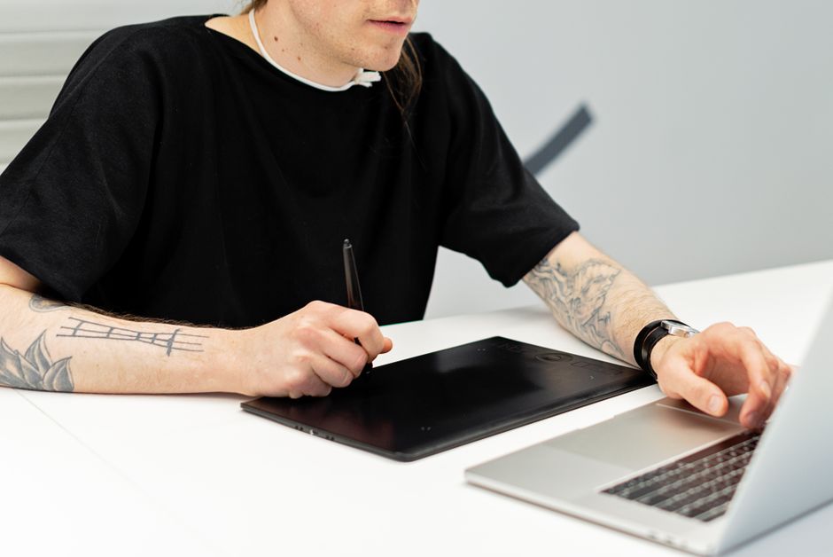 A tattooed artist working on a graphics tablet while using a laptop indoors.