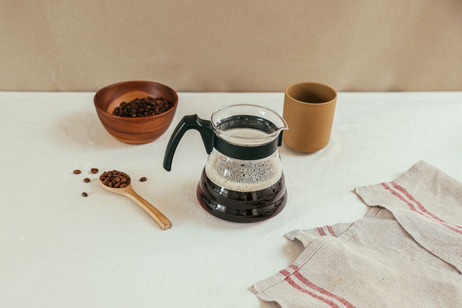 A drip coffee setup featuring a coffee pot, beans, a wooden spoon, and a cup on a white table.