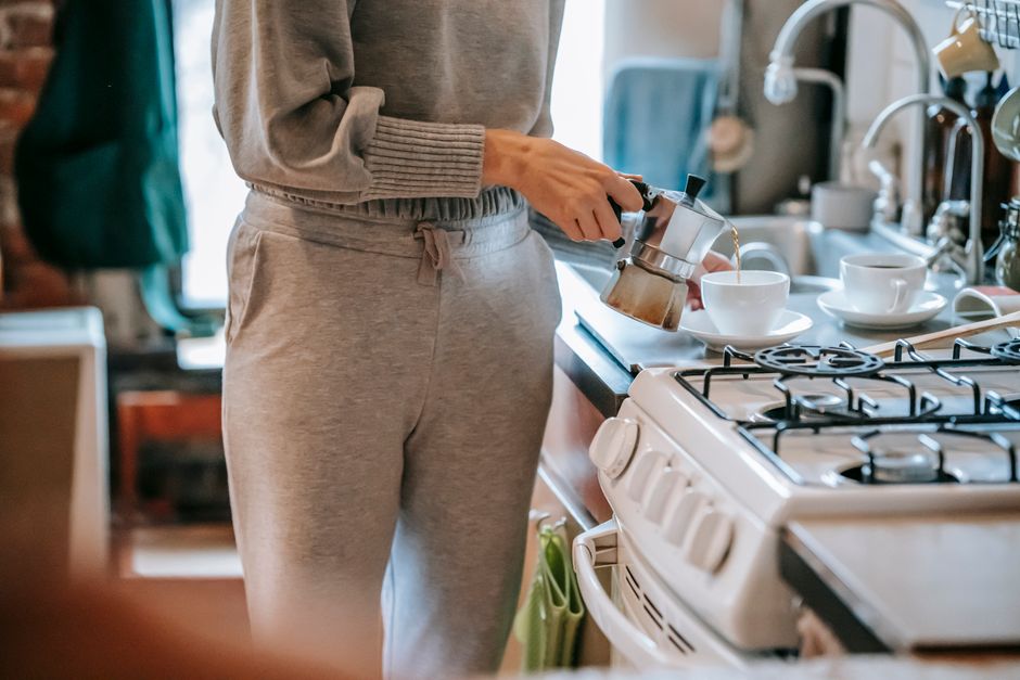Crop unrecognizable female in casual clothes pouring fresh brewed coffee form moka pot into cup while standing near stove in kitchen