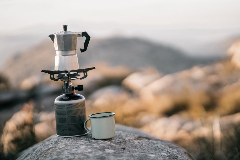 A coffee maker and cup sit atop a camping stove, set on a rock with a blurred mountain view in the background.