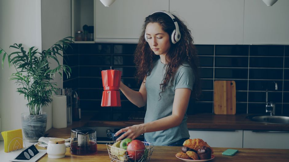 Woman in kitchen making coffee while listening to music, surrounded by fruit and pastries.