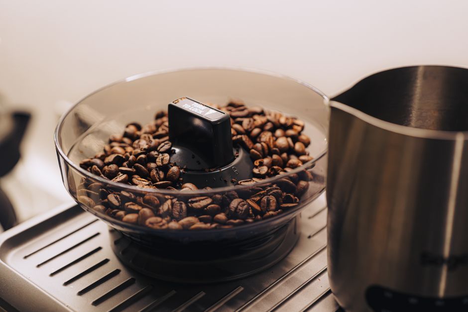 Close-up of coffee beans in a grinder beside a stainless steel milk frothing pitcher.