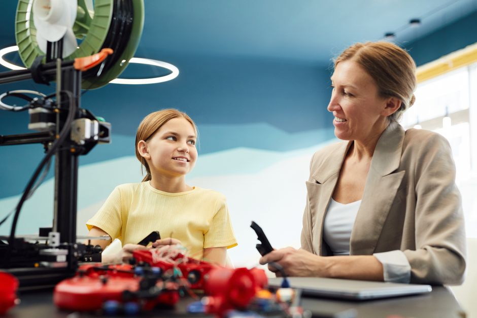 Low angle of smiling woman and little girl sitting at table and working on engineering project together while looking at each other in light workspace