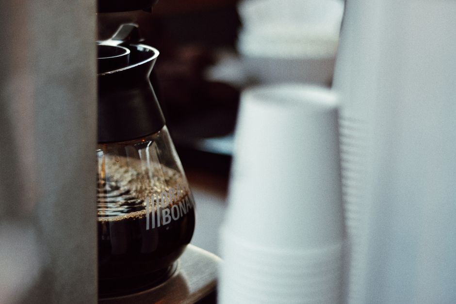 A close-up view of a coffee pot next to a stack of disposable cups in an indoor setting.