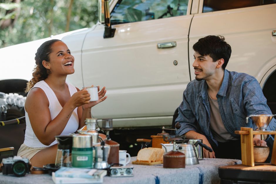 Happy young multiracial couple smiling and chilling while drinking coffee during road trip in nature on sunny day