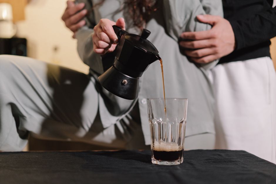 Close-up of a couple pouring freshly brewed coffee from a moka pot into a glass cup.