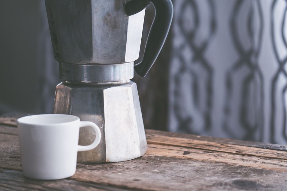 A vintage moka pot next to a white coffee cup on a rustic wooden table.