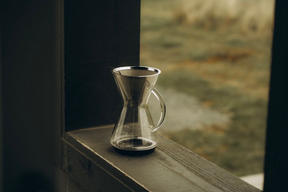 Glass pour over coffee maker placed on a wooden ledge against a blurred nature background.