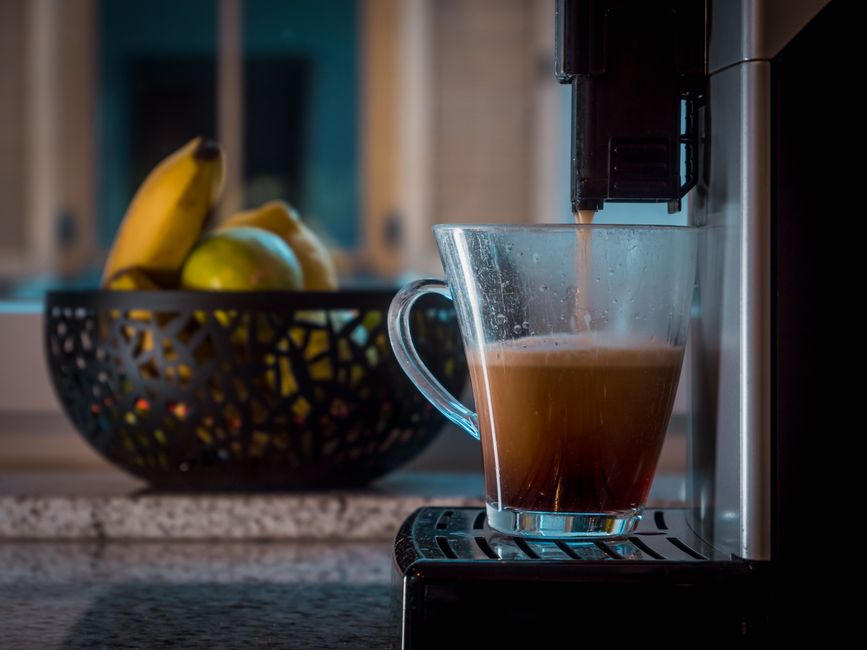 Glass cup catching freshly brewed coffee near a bowl of bananas and apples in a modern kitchen.