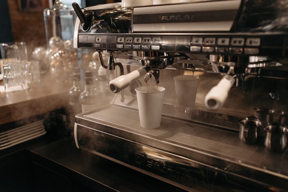 A modern espresso machine brewing coffee into a paper cup at a cozy cafe, surrounded by steam and equipment.