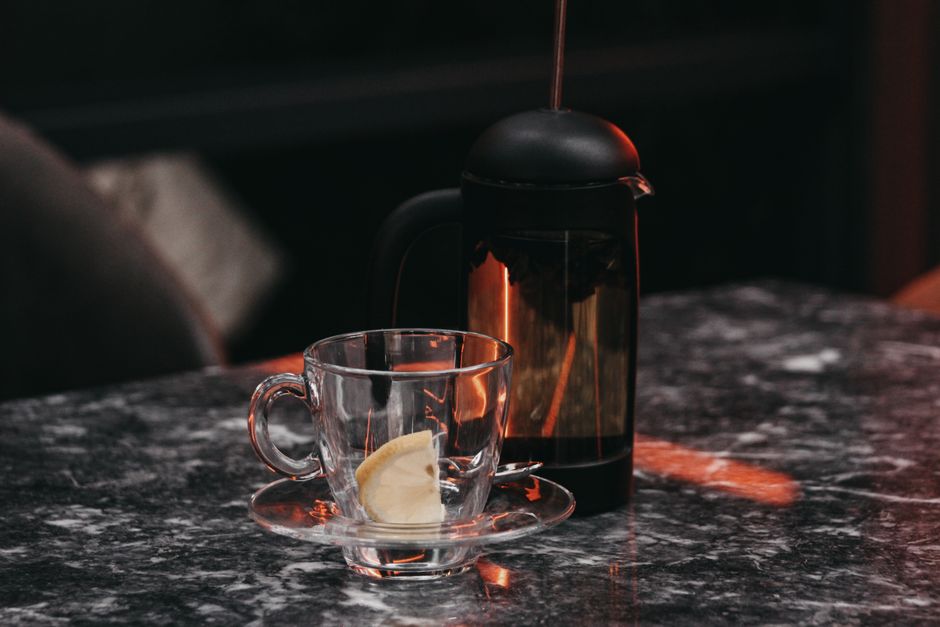 A transparent glass cup with a lemon slice and a French press on a marble table setting.