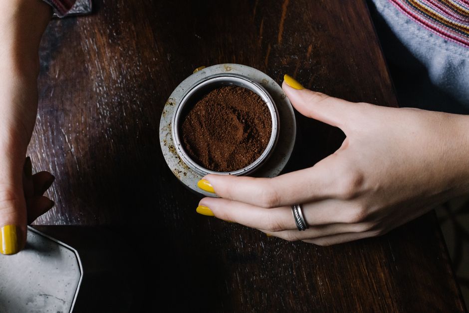 A woman's hands with yellow nails holding coffee grounds in a metal cup on a wooden table.