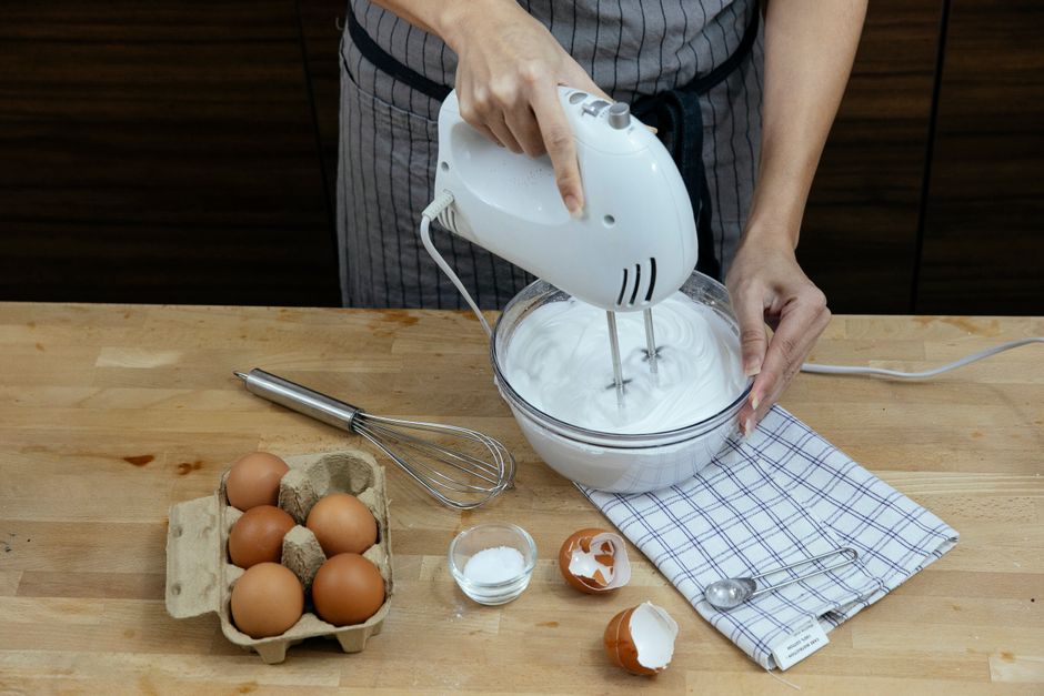 High angle crop anonymous female chef in apron beating eggs and preparing fluffy whipped cream in bowl while cooking in light kitchen