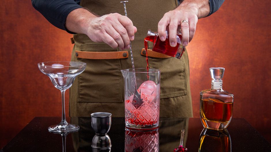 A bartender mixes a classic cocktail with red liquor, ice, and stirring implements.