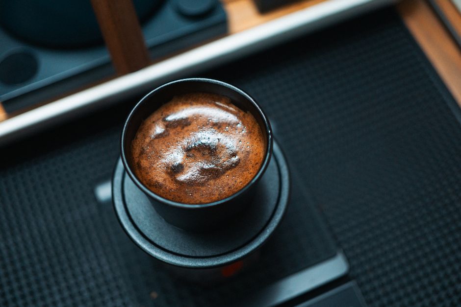 A detailed high-angle view of a freshly brewed cup of coffee with a frothy surface, on a black mat.