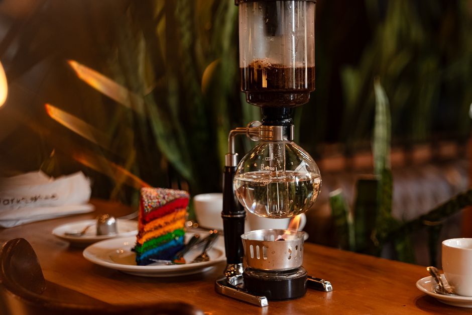 Close-up of a siphon coffee maker and colorful rainbow cake slice on a wooden table, captured indoors.